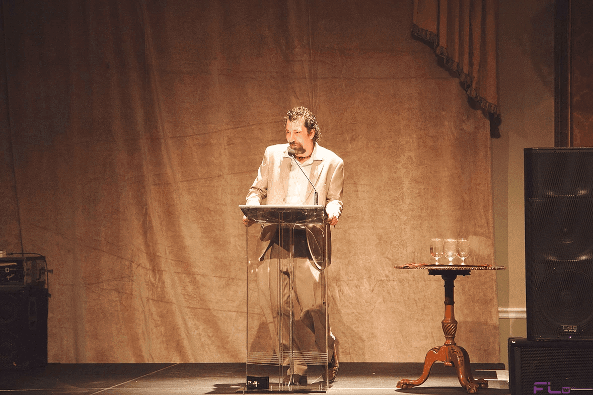 Speaker at a clear acrylic podium on stage, speaker cabinet at right, velour drape behind