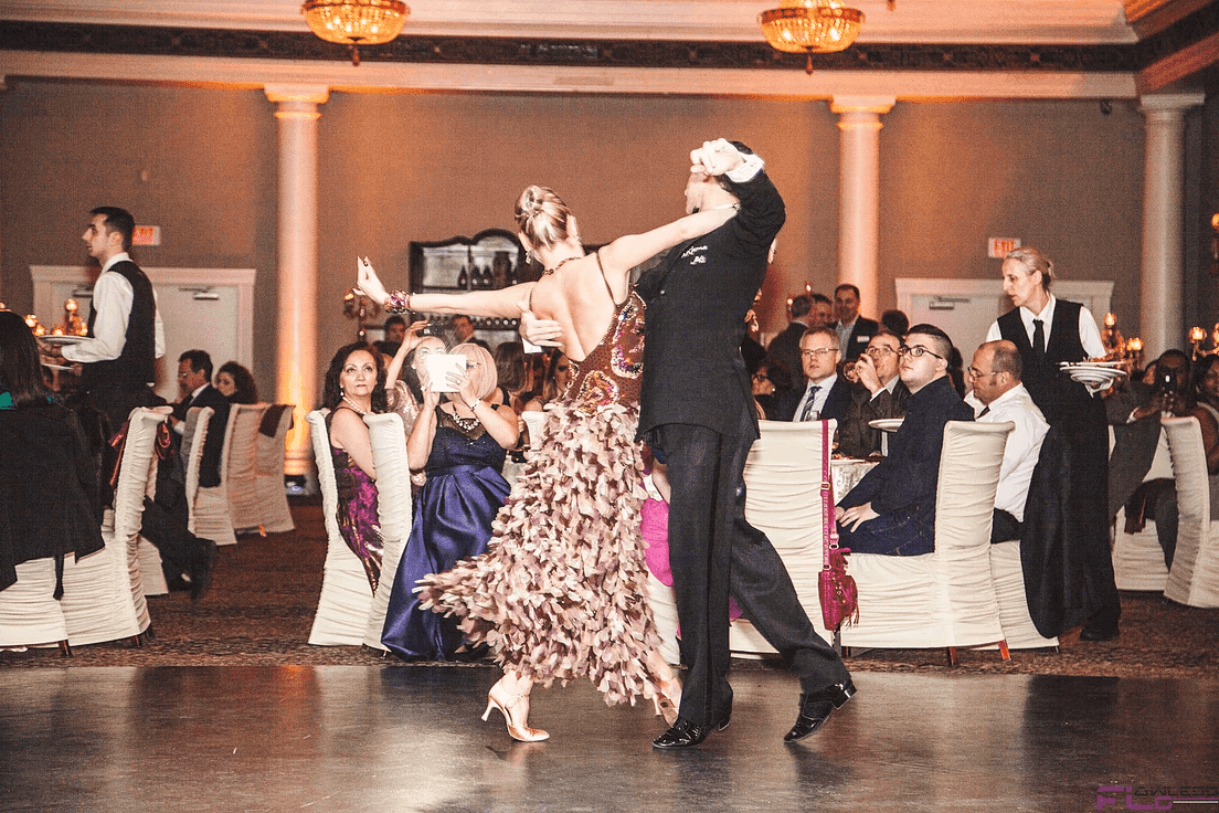 Ballroom dance couple performing among dinner rounds under amber-lit columns
