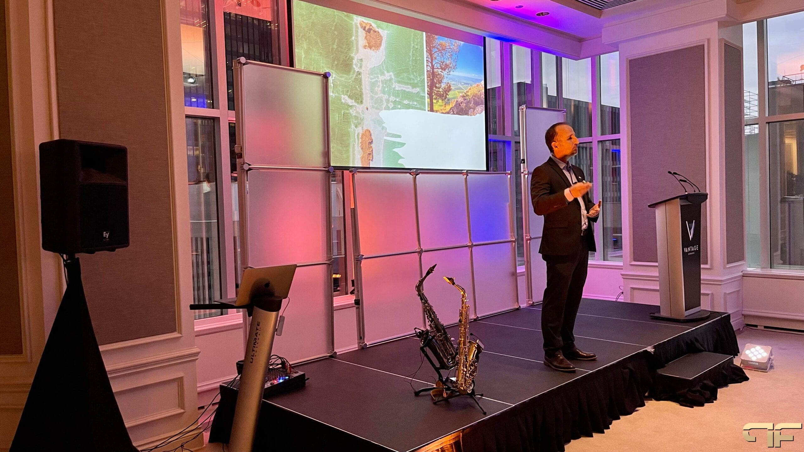 Presenter on stage beside saxophones, speaker on tripod and laptop stand at stage left