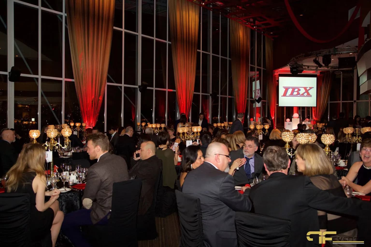 Guests at round tables during a corporate gala dinner with red uplit drapes, crystal centrepieces, and a projection screen